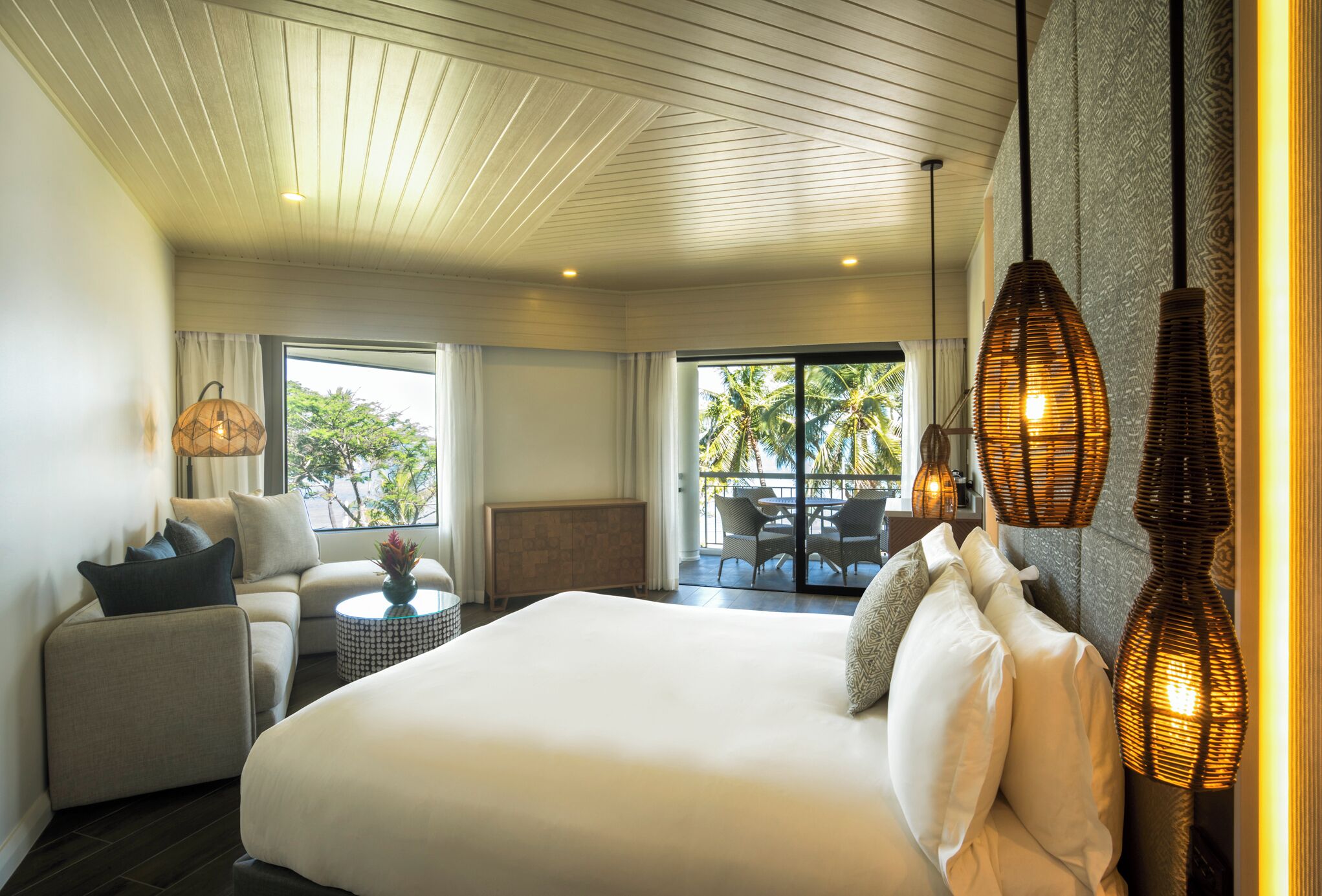A hotel room with a large bed, a sofa, a small table, and a balcony with outdoor furniture.