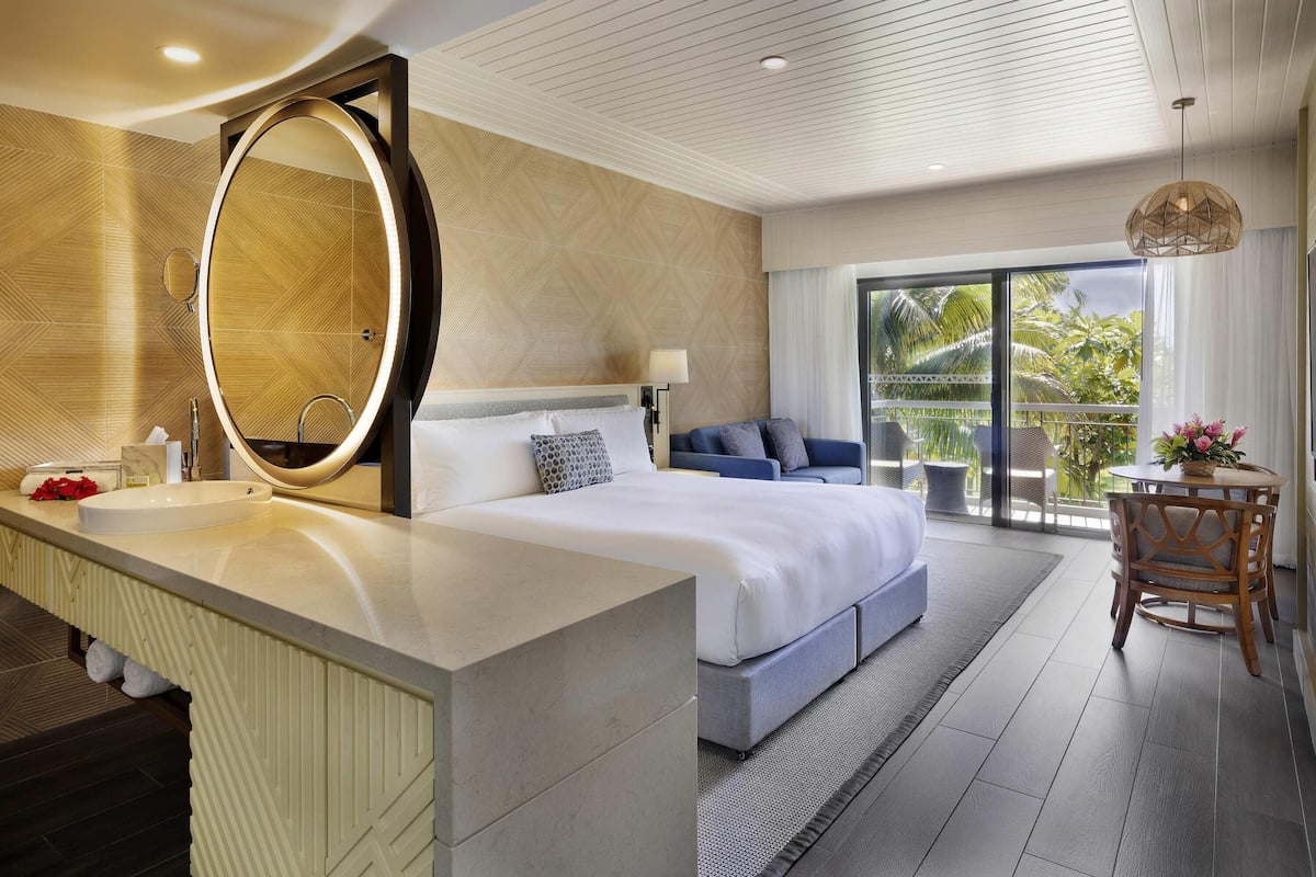 Une chambre d’hôtel moderne avec un grand lit, un miroir rond, un balcon avec vue et une petite table agrémentée de fleurs.