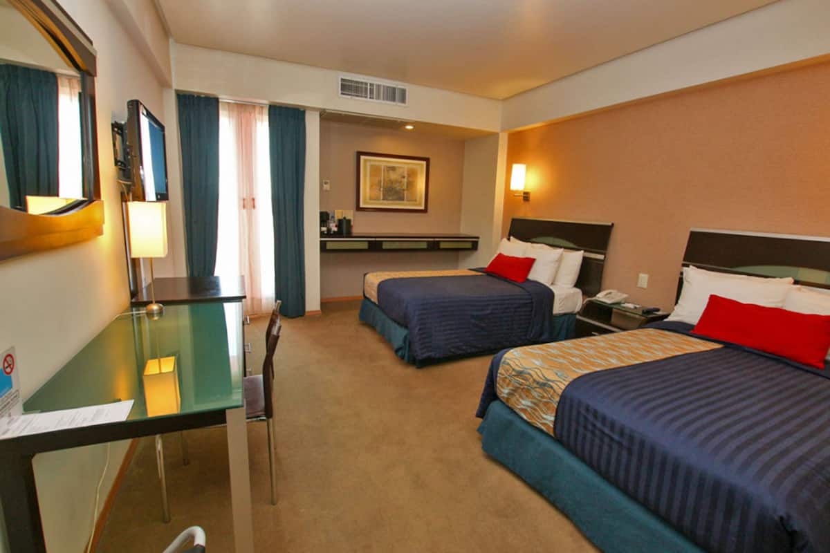 Ein Hotelzimmer mit zwei Betten, einem Schreibtisch, einem Sessel, einem Spiegel, einem Fernseher und einer kleinen Küchenzeile.