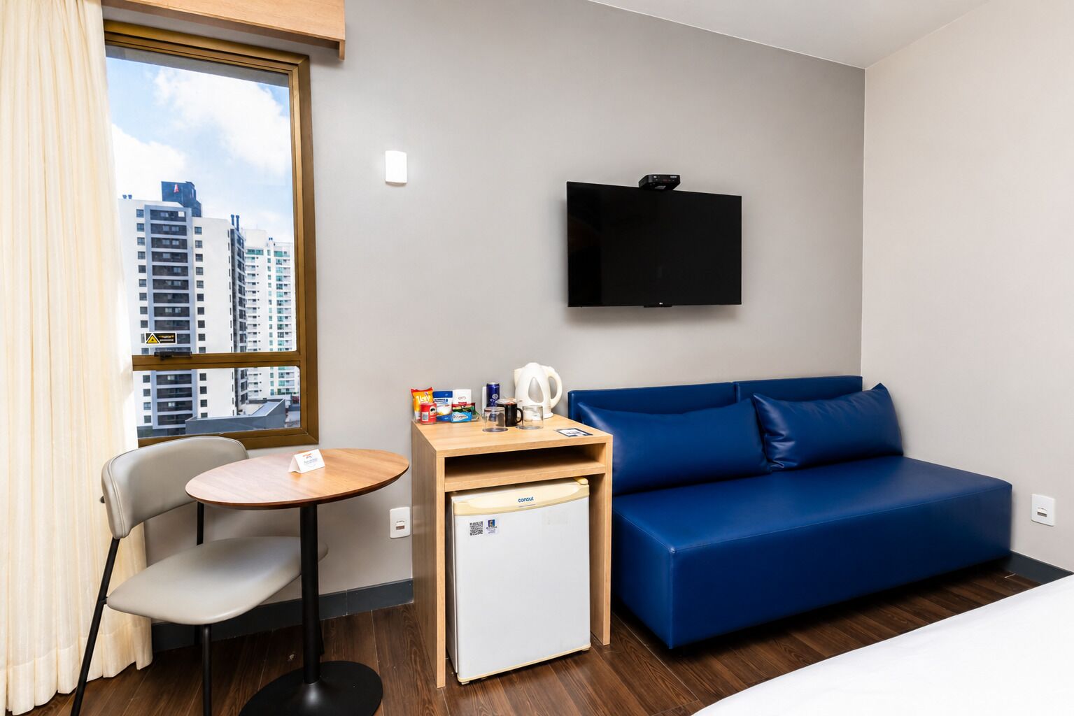 Quarto de hotel com sofá azul, uma pequena mesa com cafeteira, mini-frigorífico, televisão na parede e janela com vista para a cidade.