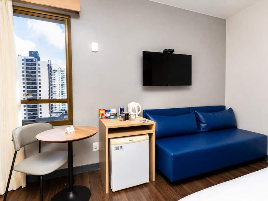 A hotel room with a blue sofa, a small table with a coffee machine, a mini fridge, a TV mounted on the wall, and a window with a city view.