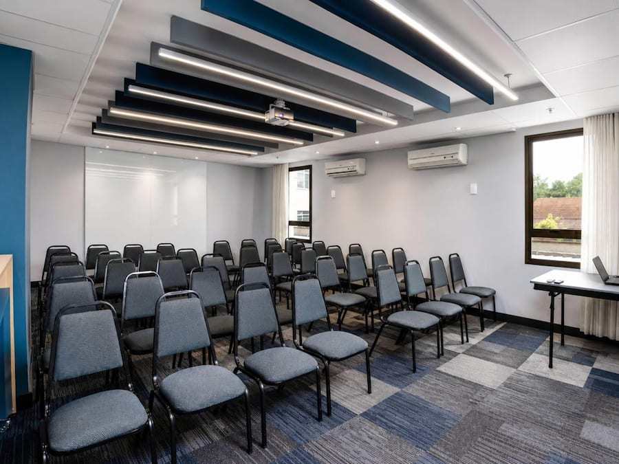 A conference room with rows of chairs, a projector, air conditioning units, and a window with curtains.