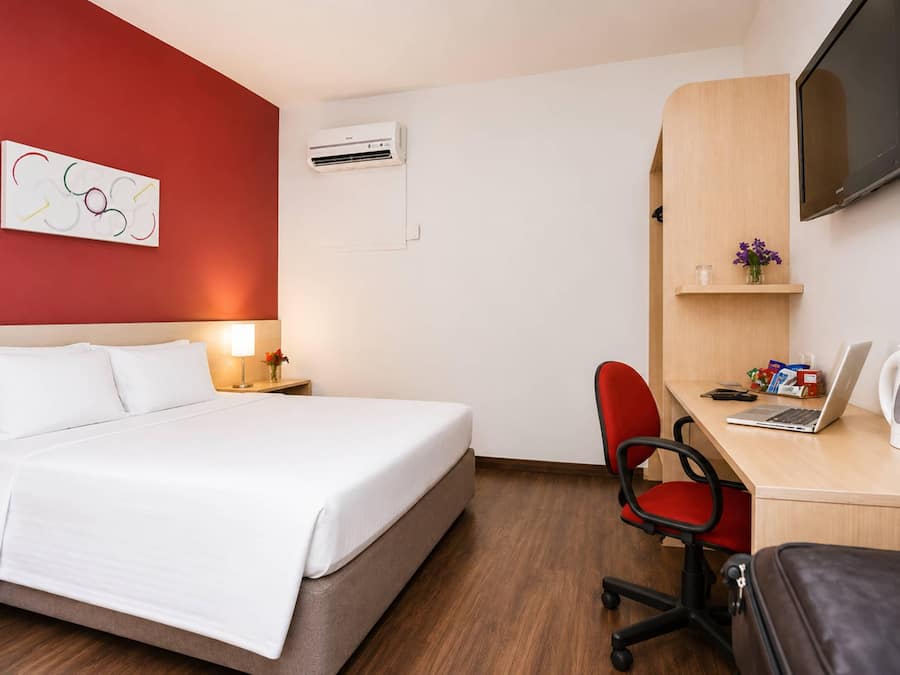 A hotel room with a bed, a desk with a laptop, a chair, a TV, and a wall-mounted shelf with a vase of flowers.