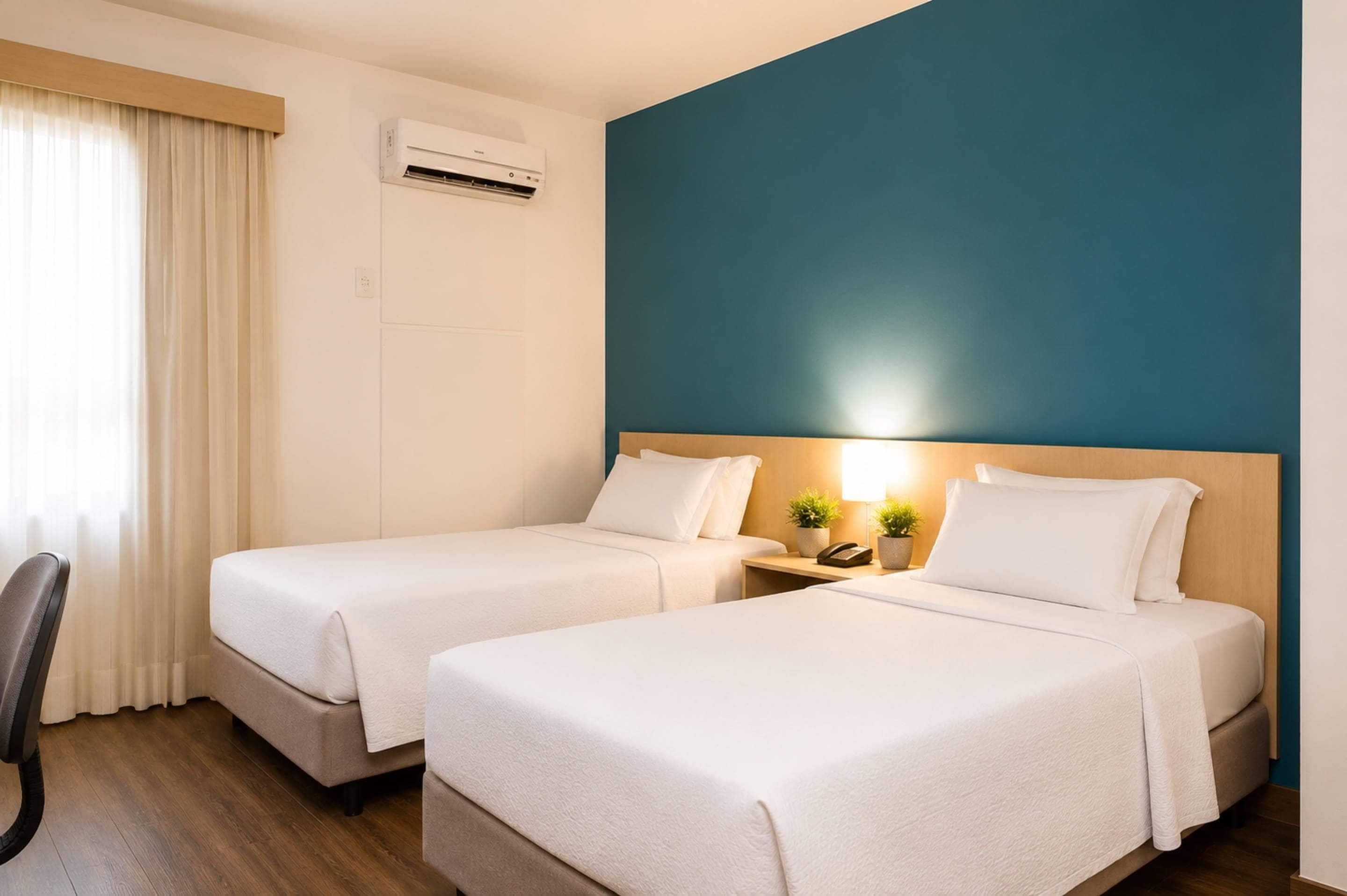 Quarto de hotel com duas camas, uma escrivaninha, aparelho de ar condicionado e vista para o exterior.