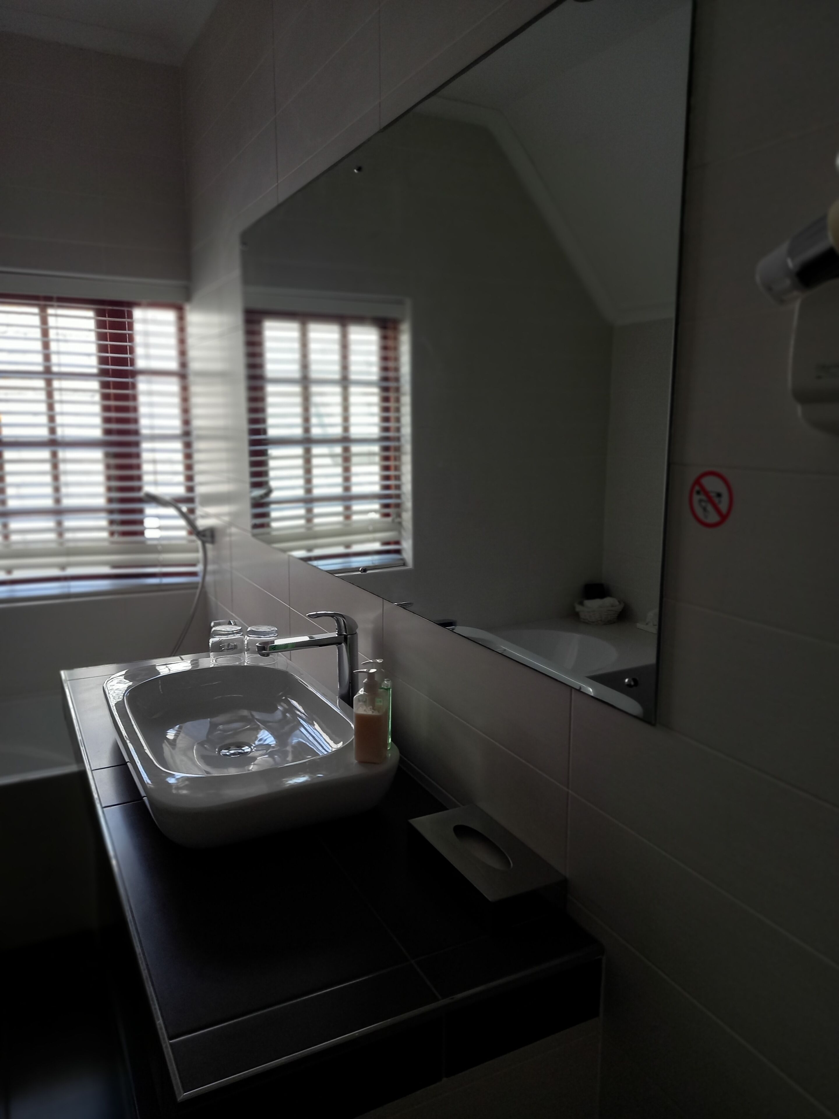Une salle de bain moderne avec un lavabo blanc, un miroir et une fenêtre avec des stores.