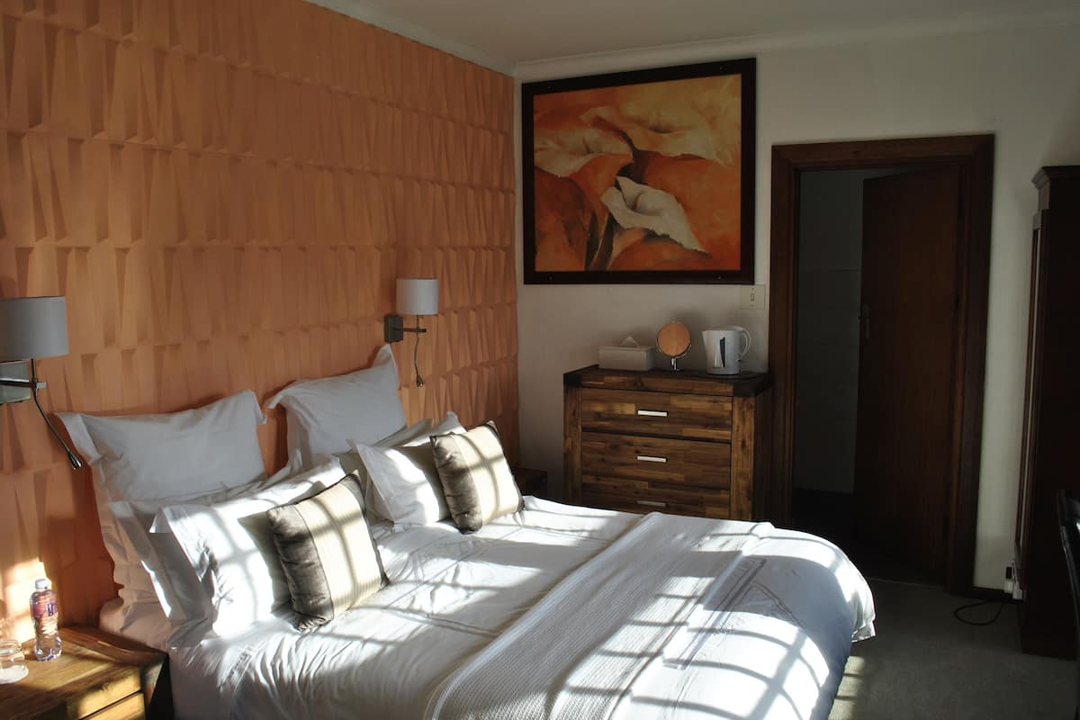 A bedroom with a wooden headboard, a bed with white bedding, a bedside table with a lamp, a dresser, and a painting on the wall.