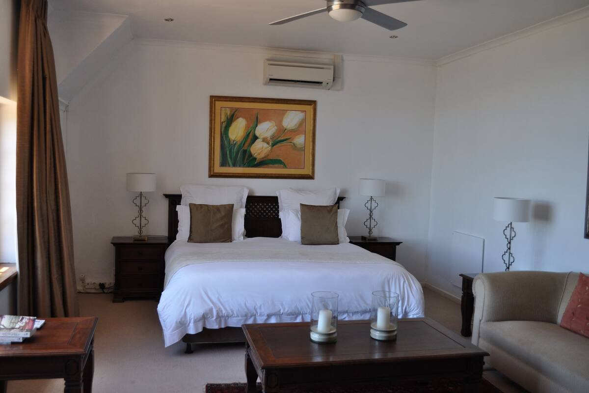 A hotel room with a large bed, two bedside tables with lamps, a ceiling fan, an air conditioning unit, and a painting of tulips on the wall.