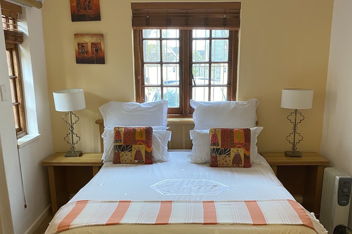 A neatly made bed with a striped bedspread, two bedside tables with lamps, a window with blinds, and two wall-mounted paintings.
