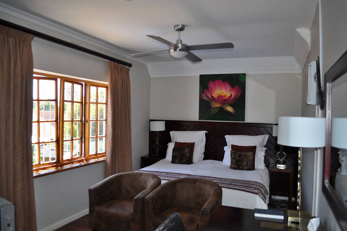 A hotel room with a bed, two chairs, a ceiling fan, a window with curtains, and a painting of a flower on the wall.