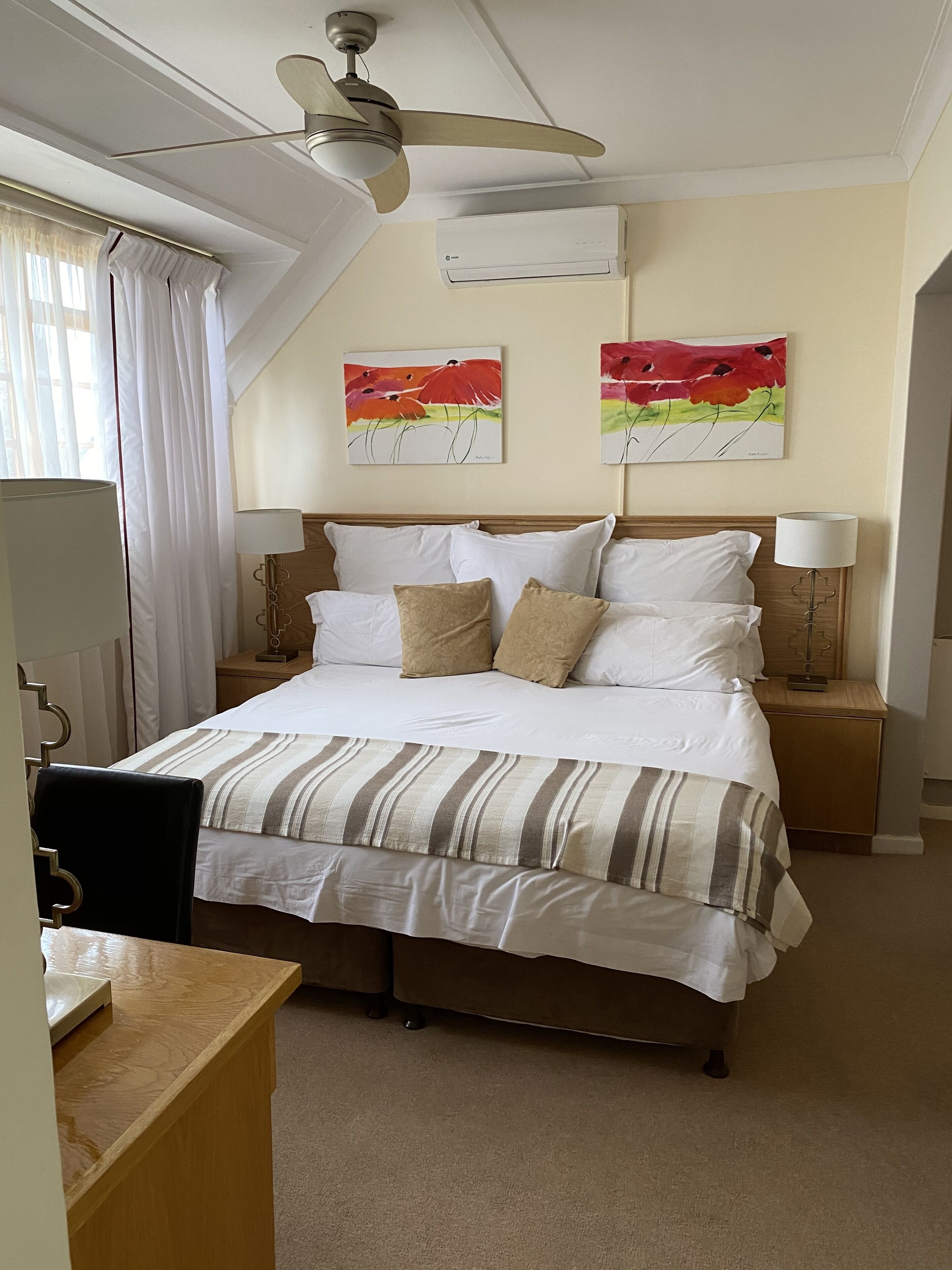 A neatly arranged hotel room with a bed, bedside lamps, a ceiling fan, and two wall-mounted air conditioning units.