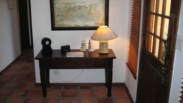 A hallway with a wooden desk, a framed painting, a lamp, and a door with window blinds.