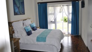 Une chambre avec un grand lit, une table de chevet en bois, un ventilateur de plafond et une vue sur l’extérieur, visible à travers une porte vitrée.
