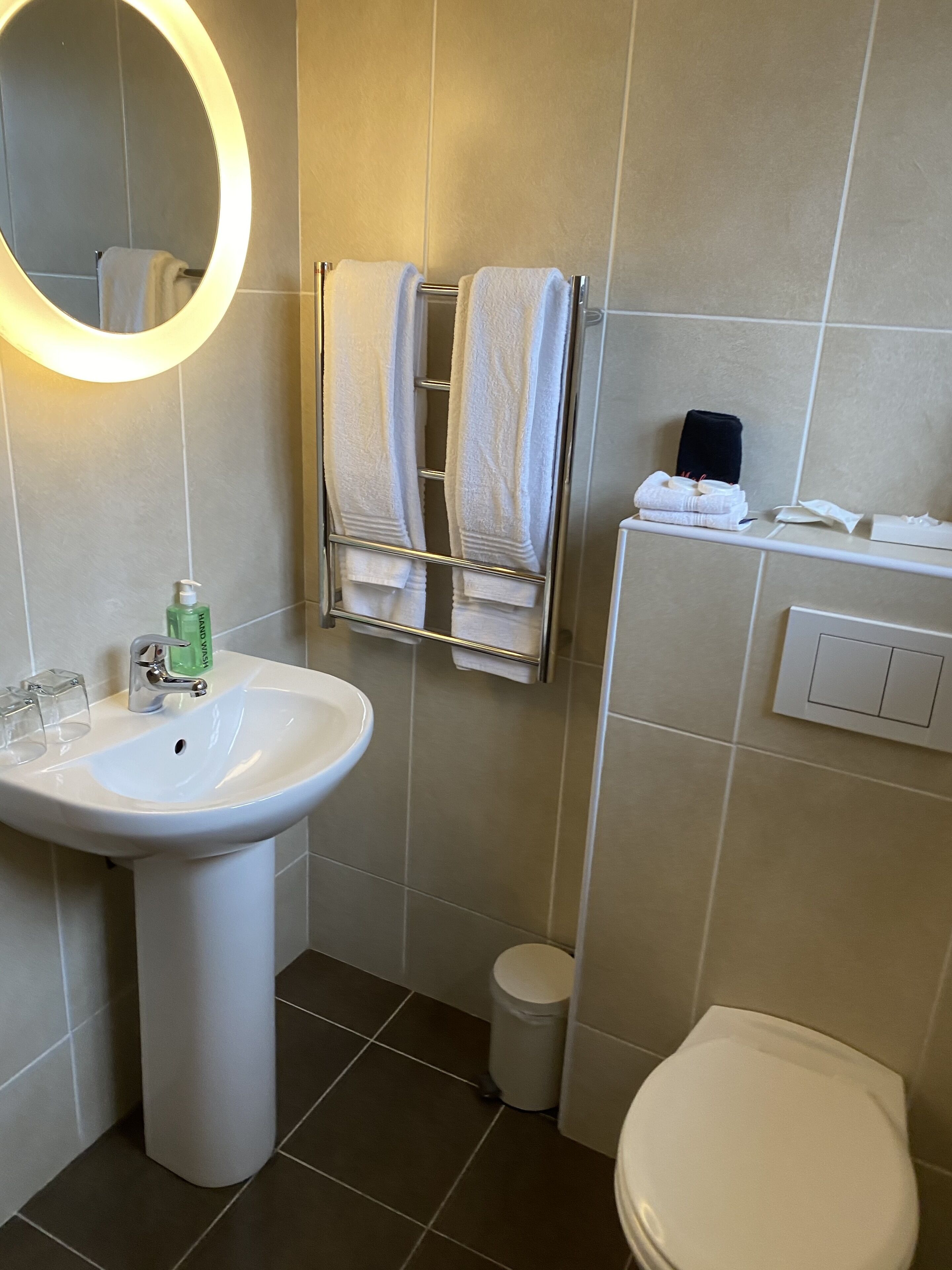A bathroom with a white sink, a mirror, two towels hanging on a rack, a toilet, and a small trash bin.