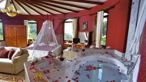 Een slaapkamer met een groot bad gevuld met rozenblaadjes, een bed met hemel en uitzicht op groen door de ramen.