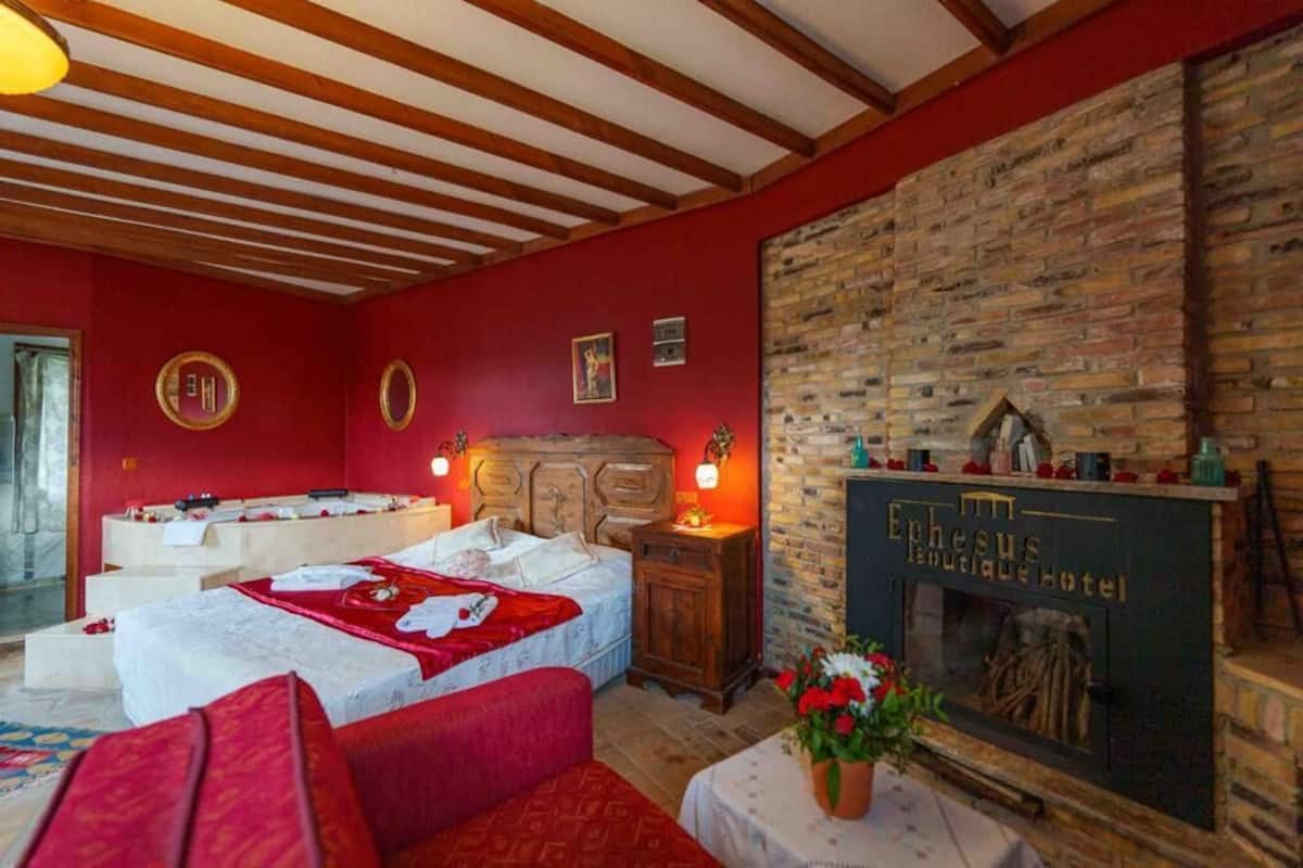 Une chambre d’hôtel avec une cheminée en pierre, un lit recouvert d’une courtepointe rouge et un plafond en bois.