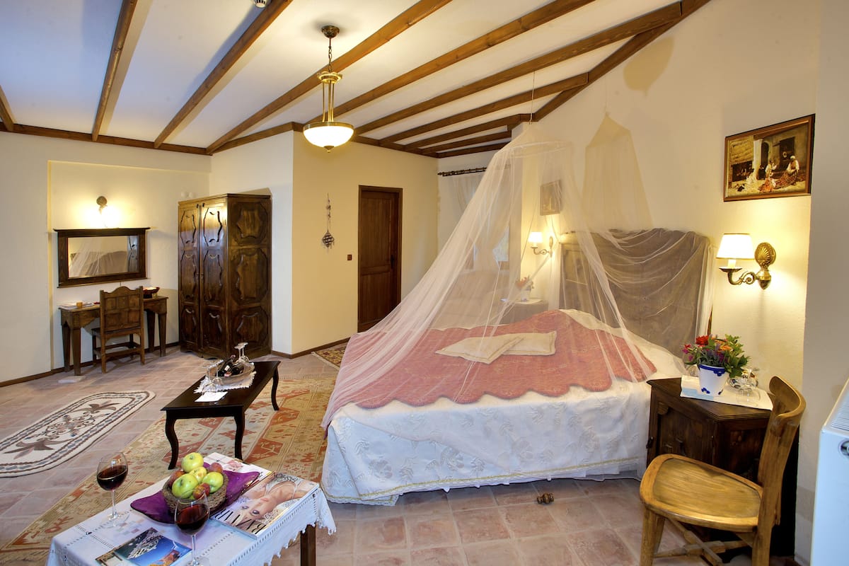 Une chambre à coucher avec un lit à baldaquin, un bureau en bois, une chaise, une table avec un magazine et un panier de pommes, et une table de chevet avec une lampe.