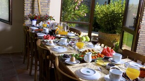 Een ontbijttafel gedekt met diverse gerechten, drankjes en fruit, met uitzicht op groen buiten.