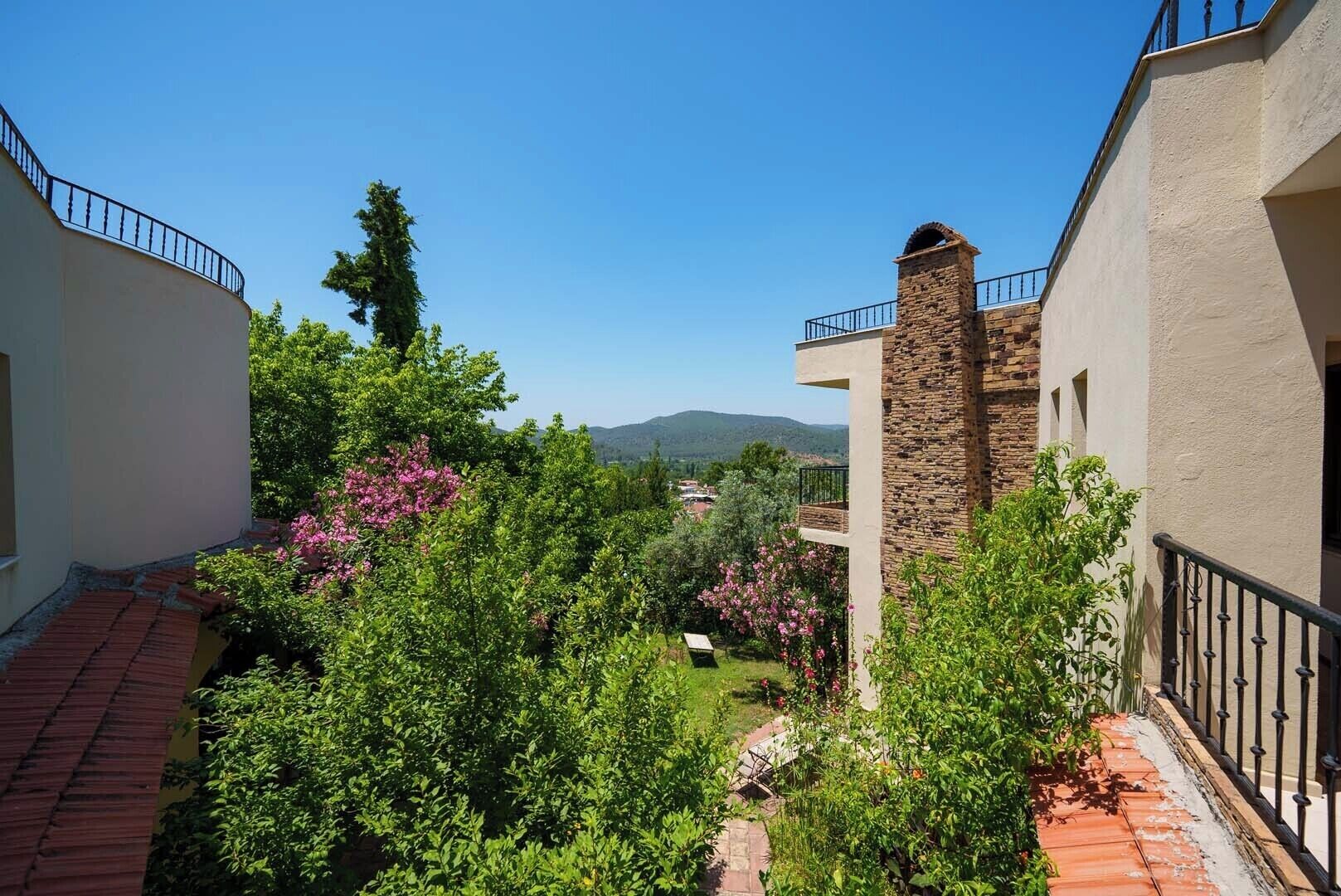 Un patio con una chimenea de piedra, tejado de tejas rojas y abundante vegetación.