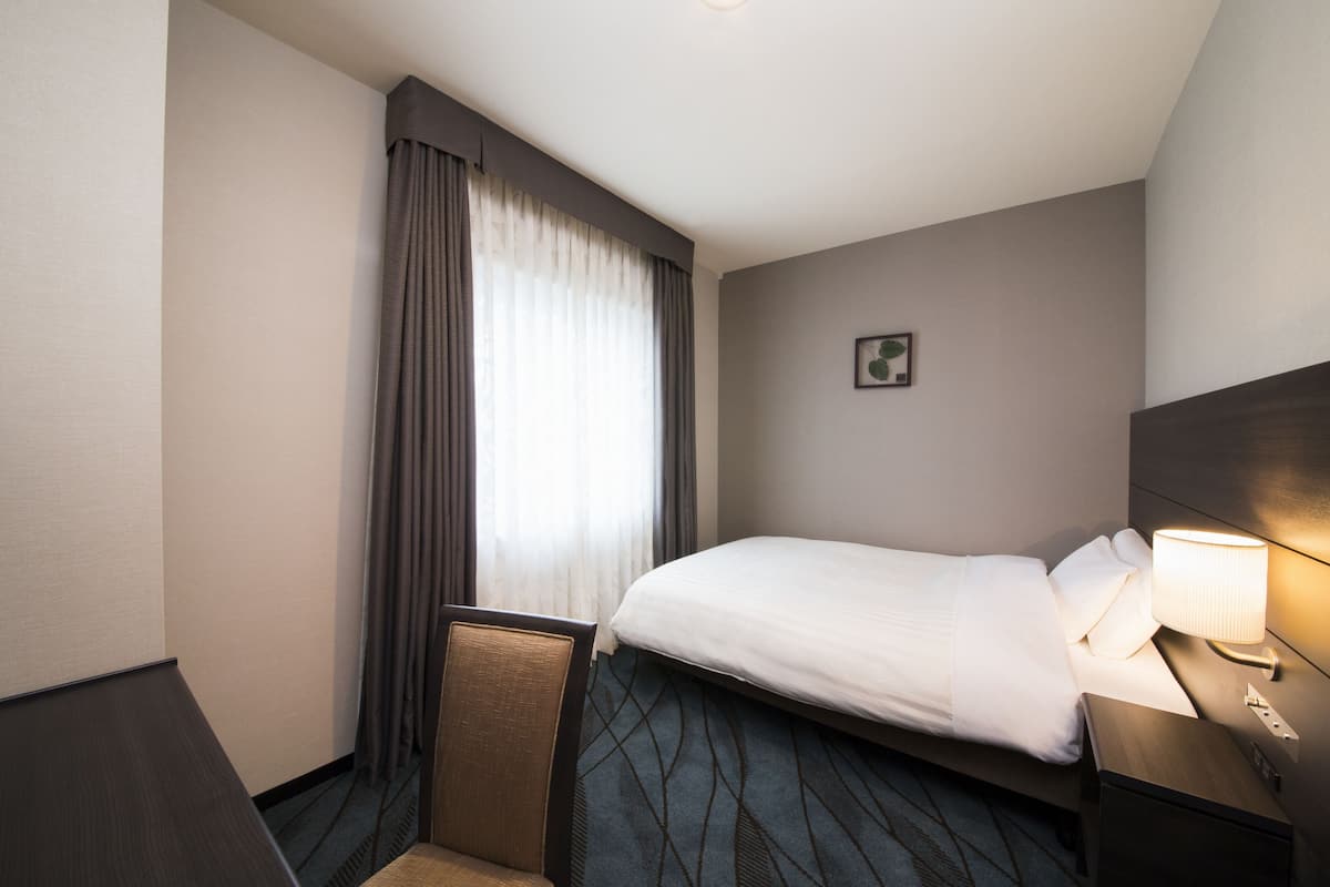 A hotel room with a bed, a chair, a nightstand with a lamp, a window with curtains, and a small picture on the wall.