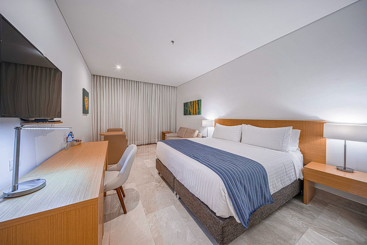 A modern hotel room with a large bed, a desk with a flat-screen TV, a chair, a bedside table with a lamp, and a large window with curtains.