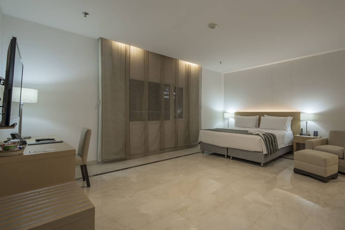 A modern hotel room with a bed, bedside tables, a desk, a chair, and a large mirror.