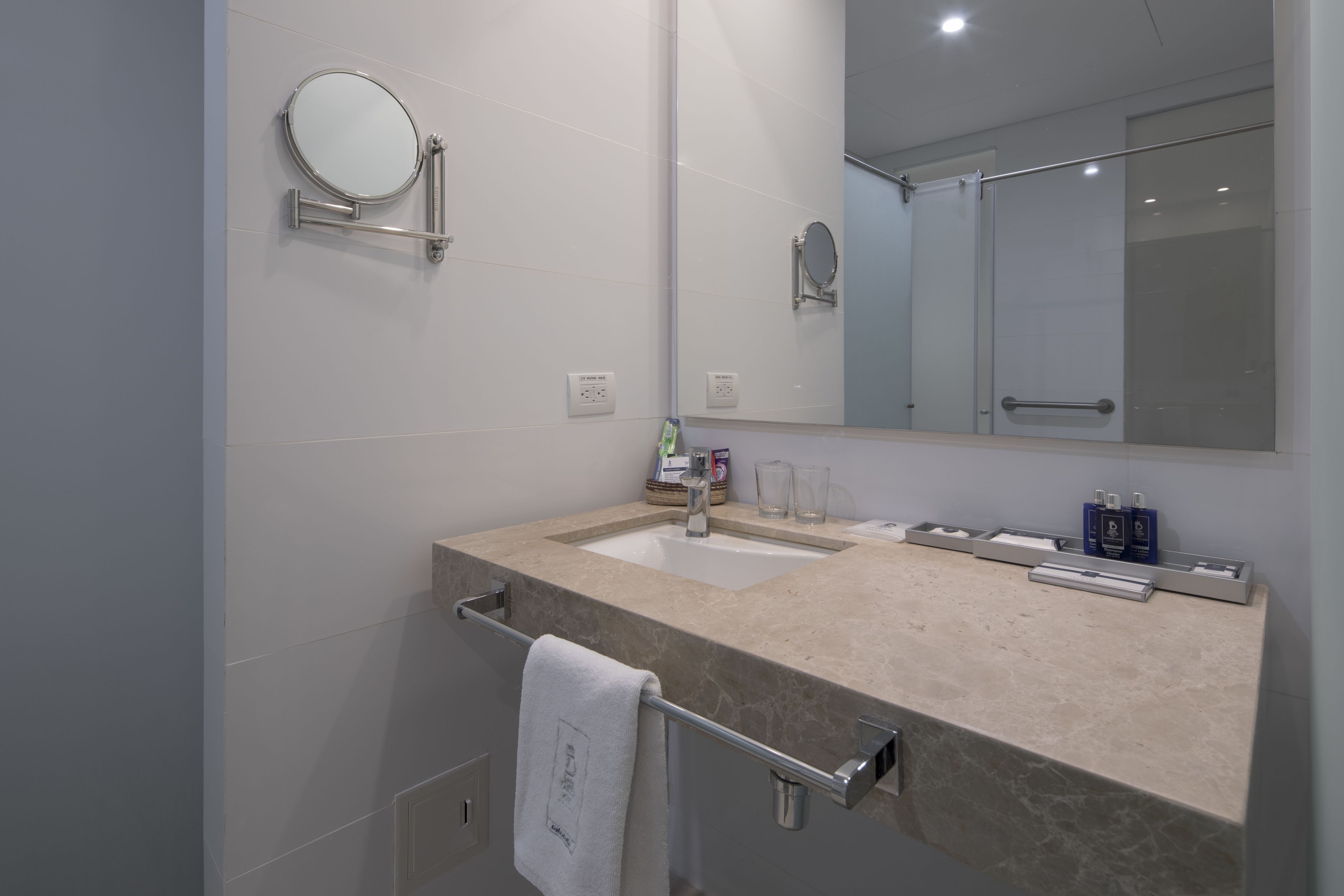 A modern bathroom with a beige marble sink, a round mirror, and a glass shower enclosure.