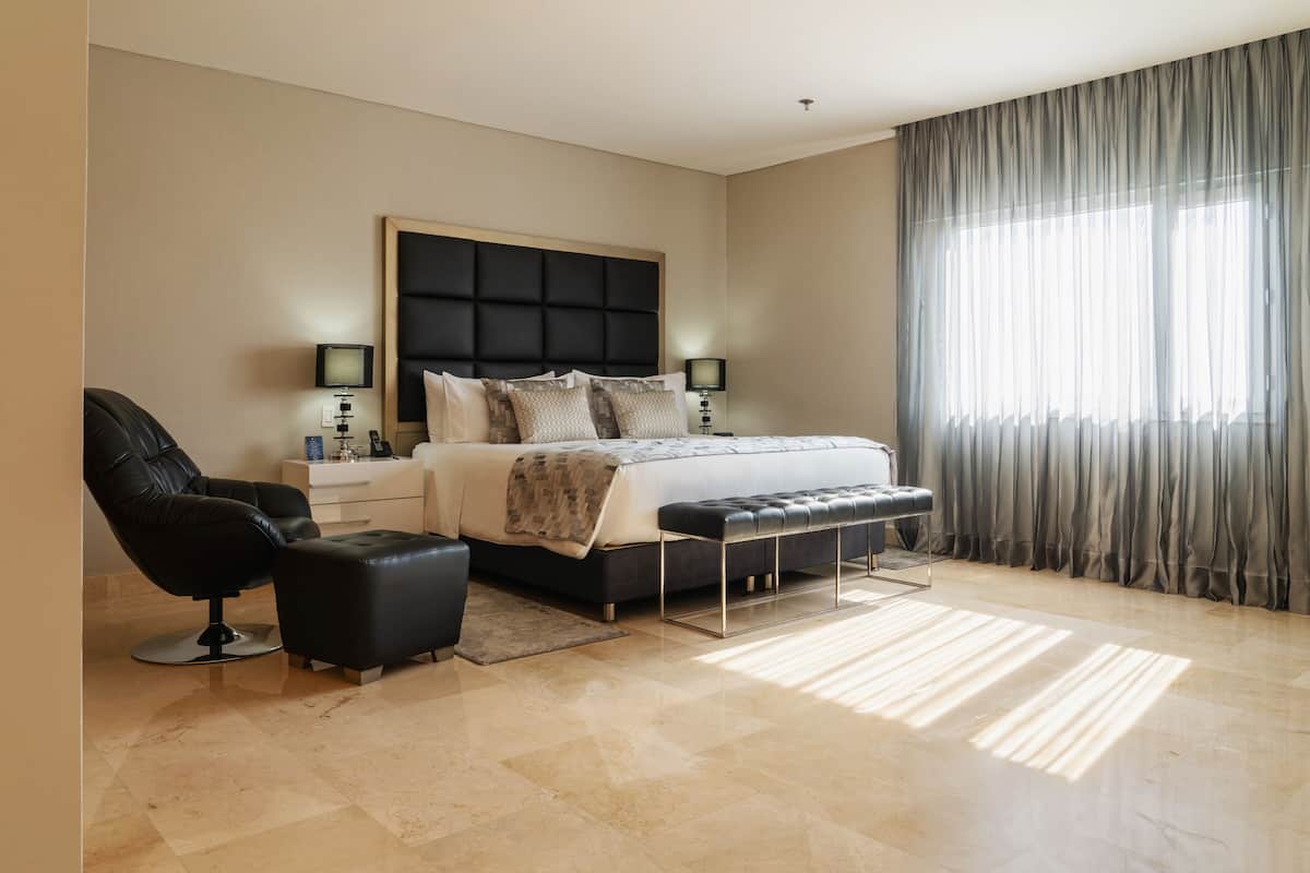 A modern bedroom with a large bed, a black leather chair, a bedside table with a lamp, and a large window with sheer curtains.