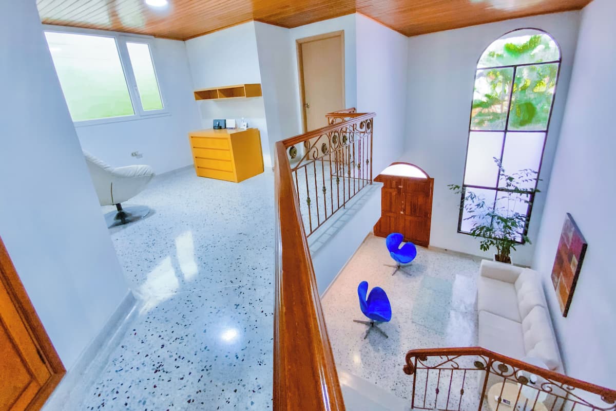 A spacious room with a wooden staircase, a white chair, a yellow dresser, and a large window with a view of greenery.