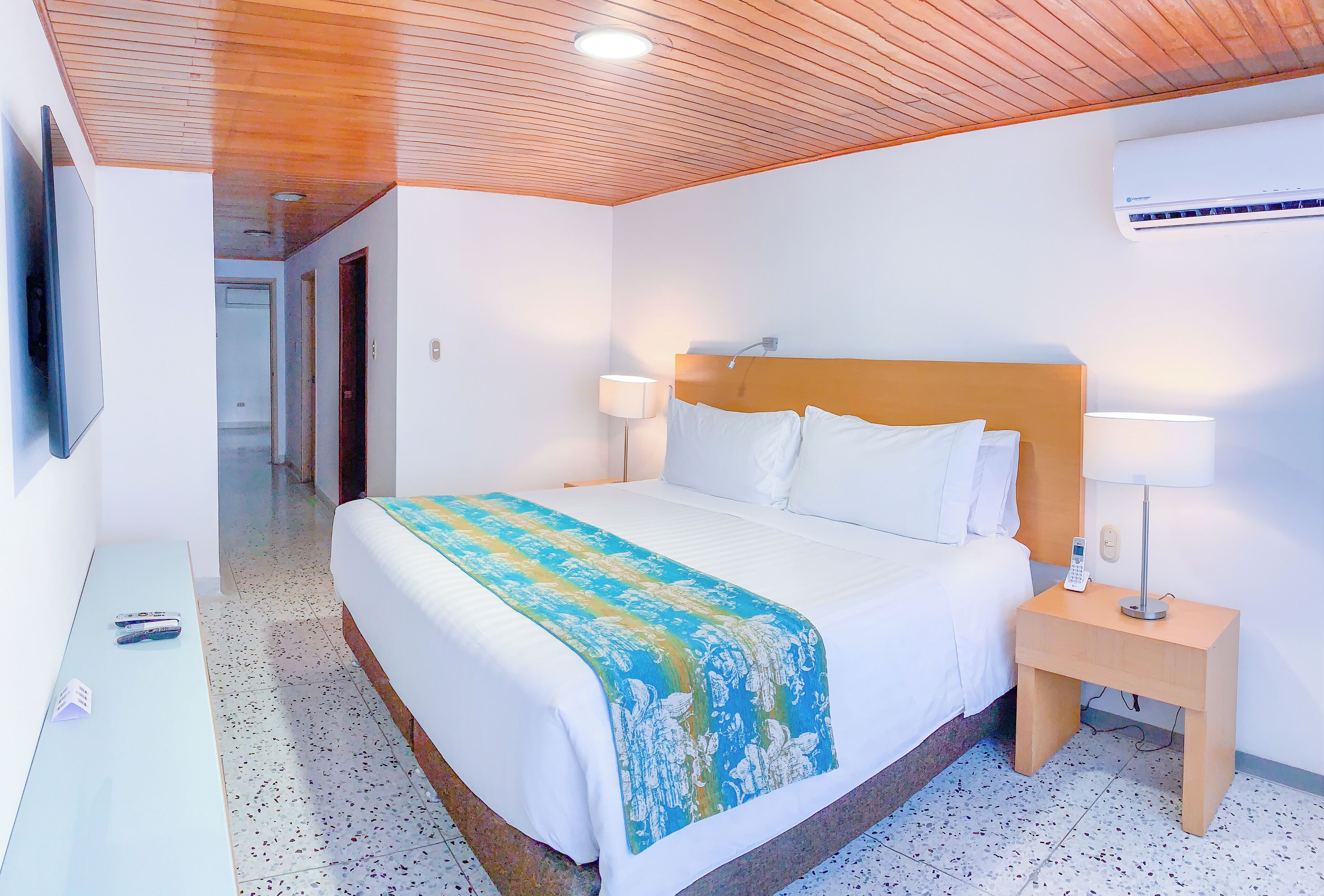 Una habitación de hotel con techo de madera, una cama con ropa blanca, una mesita de noche con lámpara y un equipo de aire acondicionado en la pared.