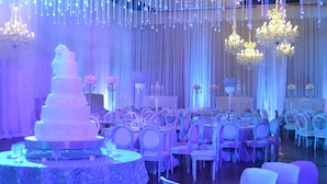 A wedding reception hall with a multi-tiered wedding cake, chandeliers, and rows of white chairs. - Dann Carlton Barranquilla (Barranquilla)