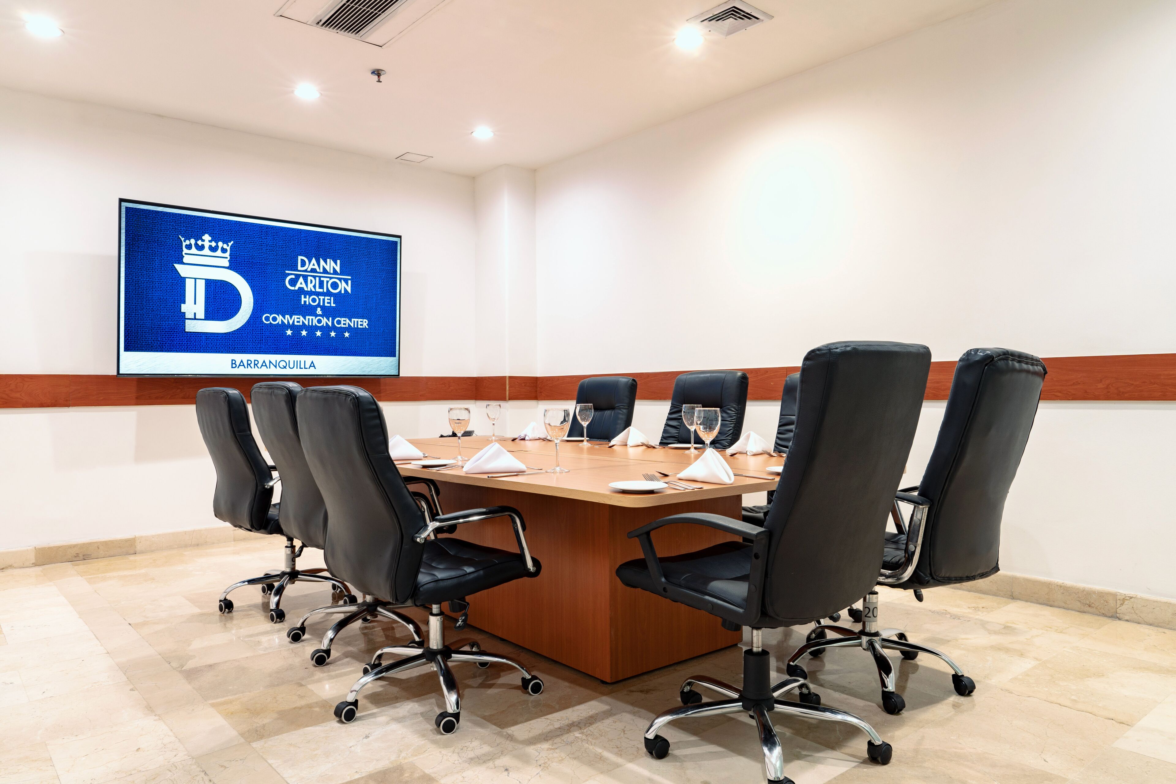 A conference room with a wooden table, black chairs, and a large screen displaying the hotel's name.