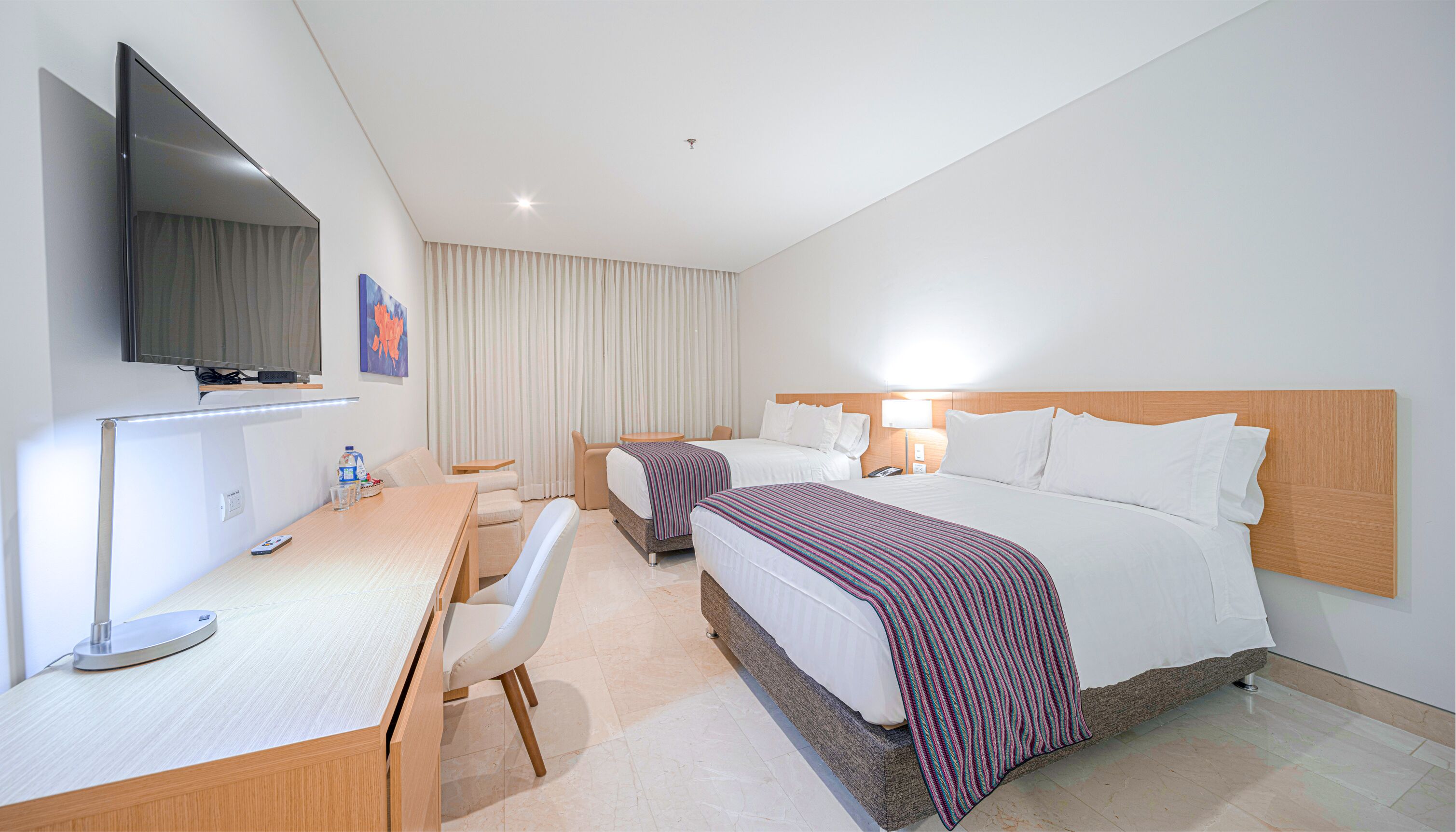 Habitación de hotel con cama, mesitas de noche, escritorio con silla, televisor en la pared y ventana con cortinas.