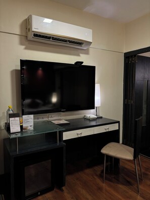 A hotel room with a large flat-screen TV, a Samsung refrigerator, a Panasonic air conditioner, a glass table with a Samsung refrigerator, a black desk with a Samsung TV, a black chair, and a wooden floor.