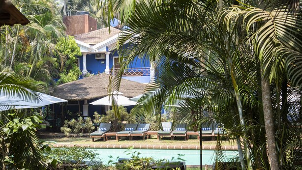 A poolside area with lounge chairs and umbrellas, surrounded by palm trees and a building with a tiled roof. - Smith & Appy Nagoa (Calangute)