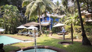 A pool area with lounge chairs and umbrellas, surrounded by palm trees and a blue building. - PRESA di GOA (Calangute)