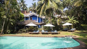 A pool area with lounge chairs and umbrellas, surrounded by palm trees and a blue building. - Smith & Appy Nagoa (Calangute)