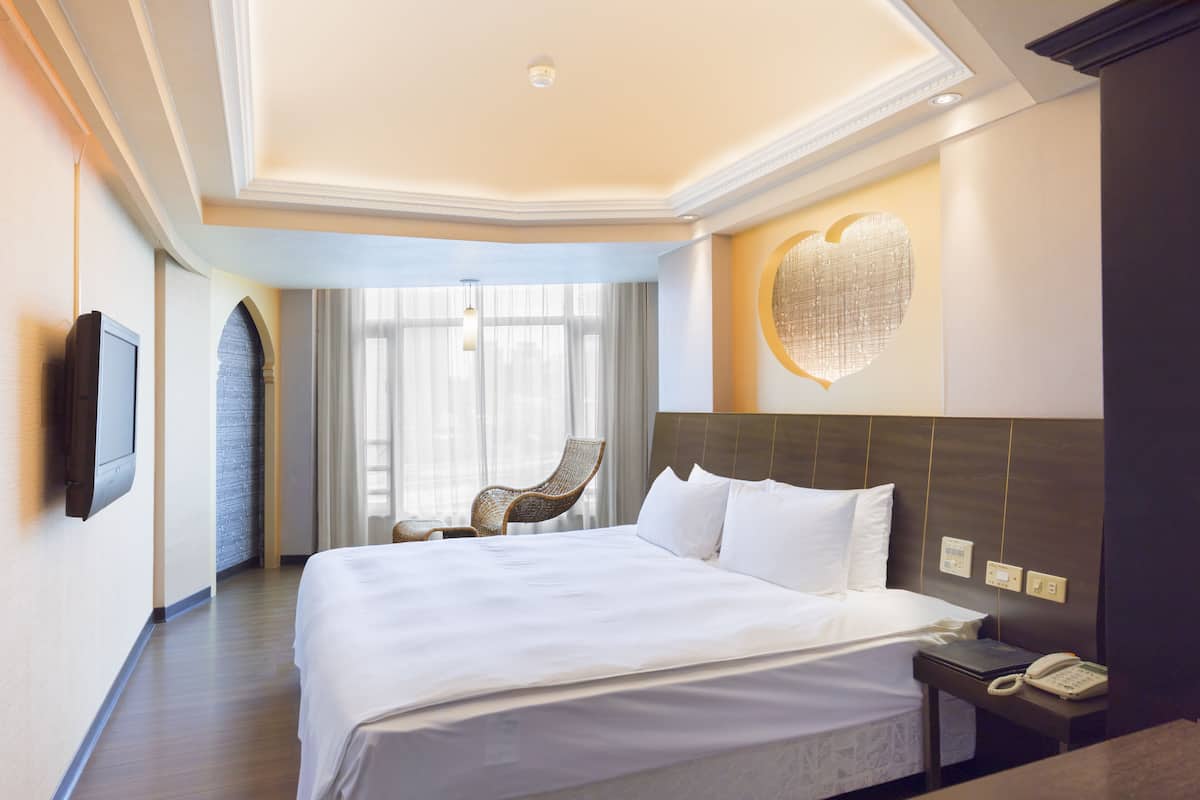 A hotel room with a large bed, a bedside table with a phone, a wall-mounted TV, a wicker chair, and a large window with curtains.