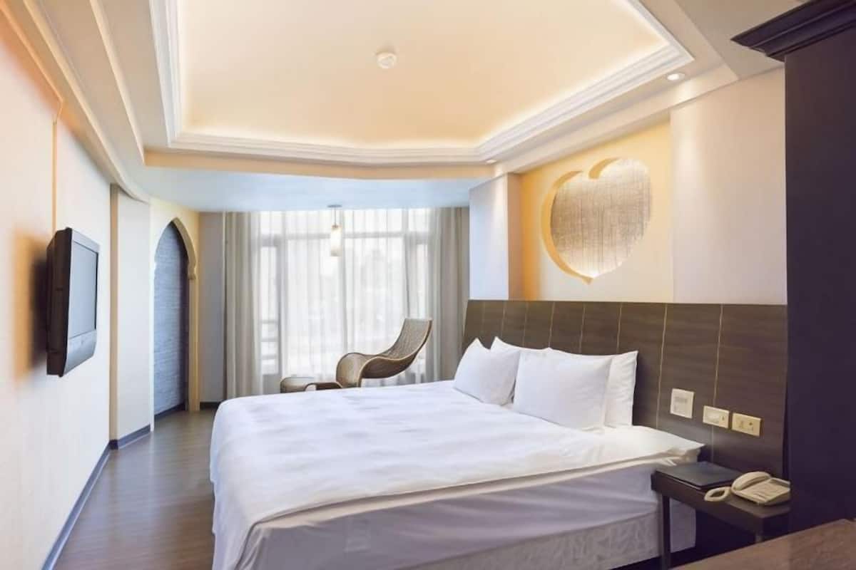 A hotel room with a large bed, a chair, a desk with a phone, and a wall-mounted TV.