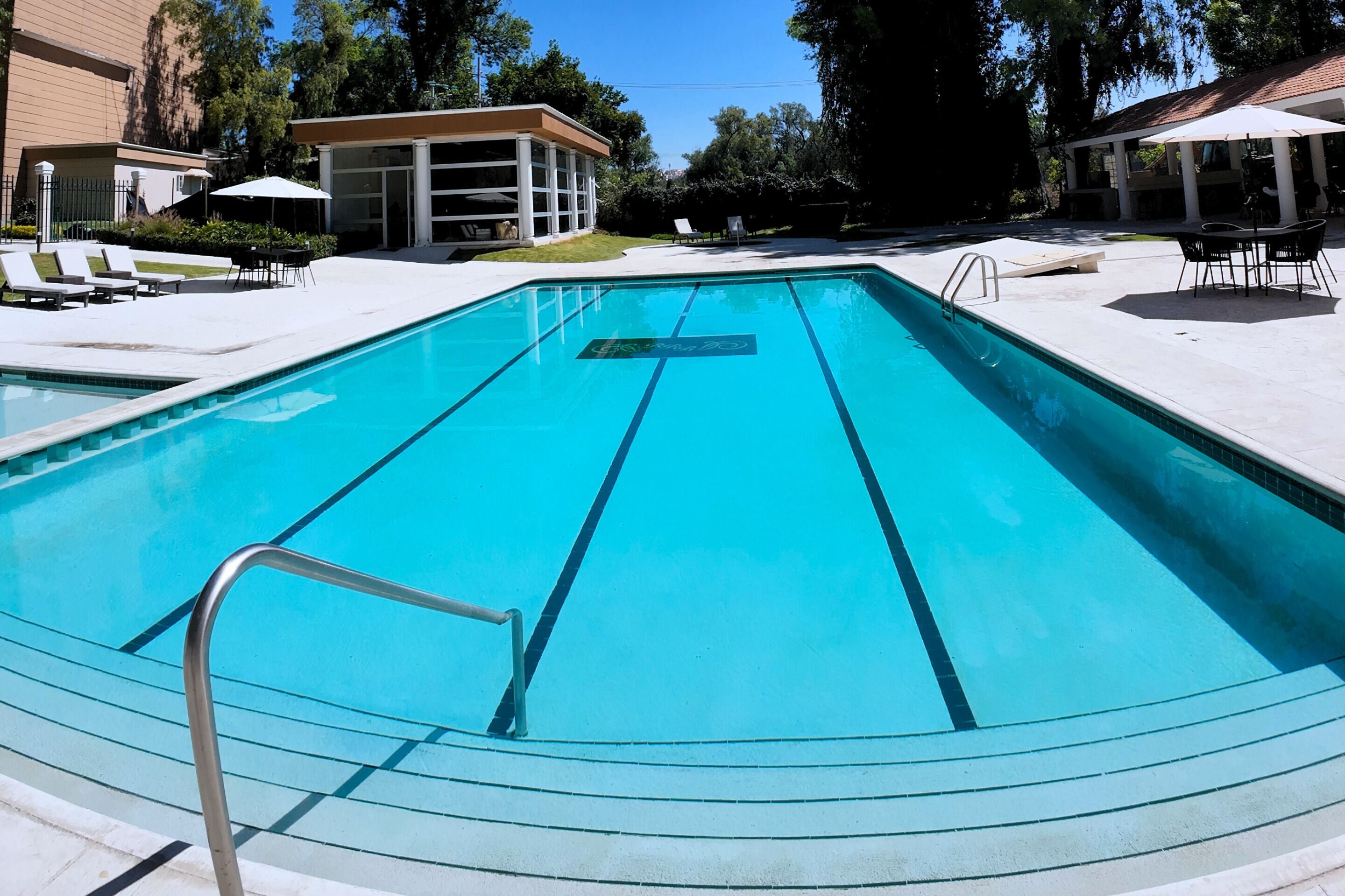 Piscina rectangular con escalera de metal, rodeada de sillones de descanso y sombrillas.