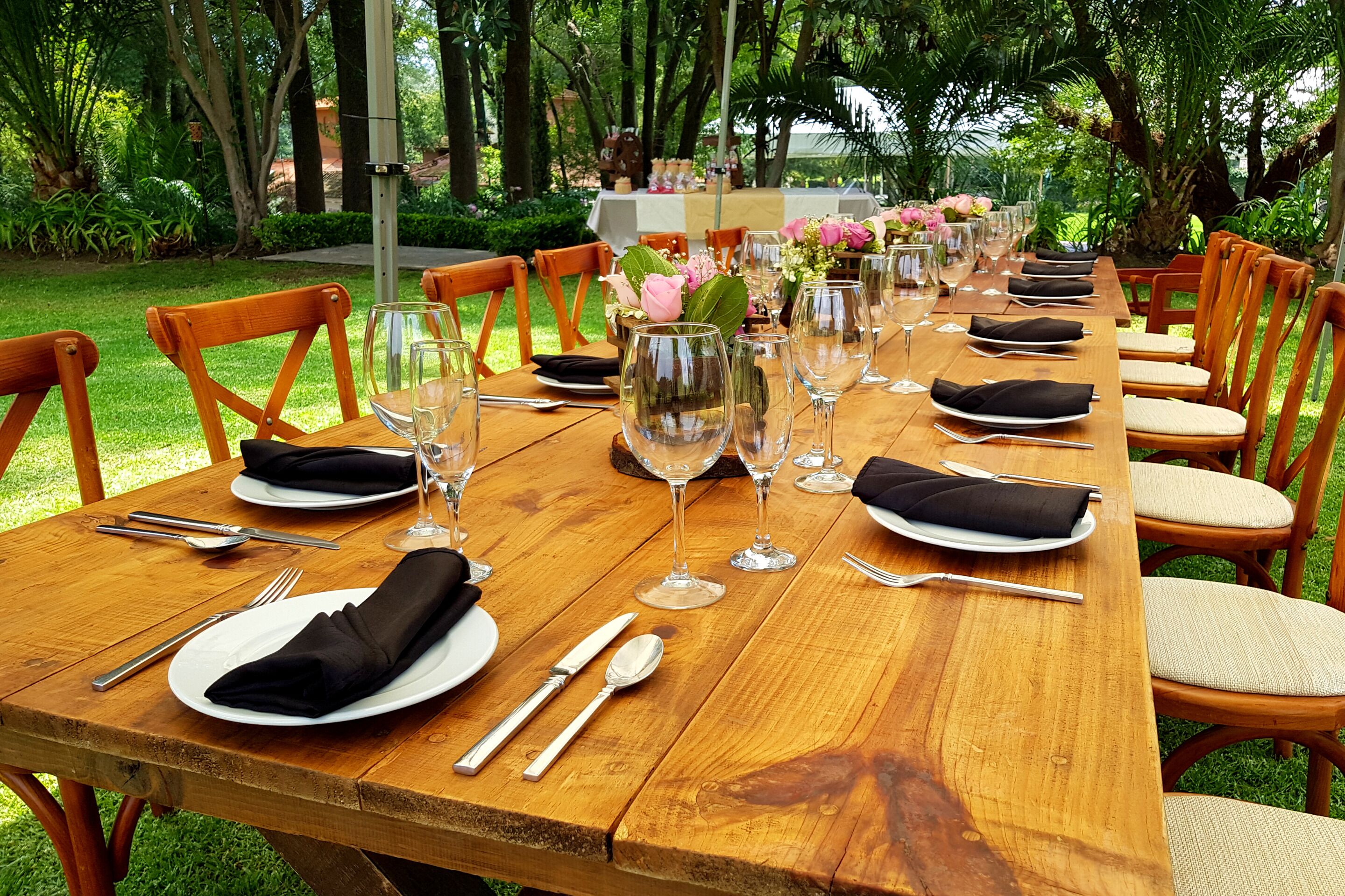 Mesa de madera con vajilla, copas de vino y sillas, lista para una comida.