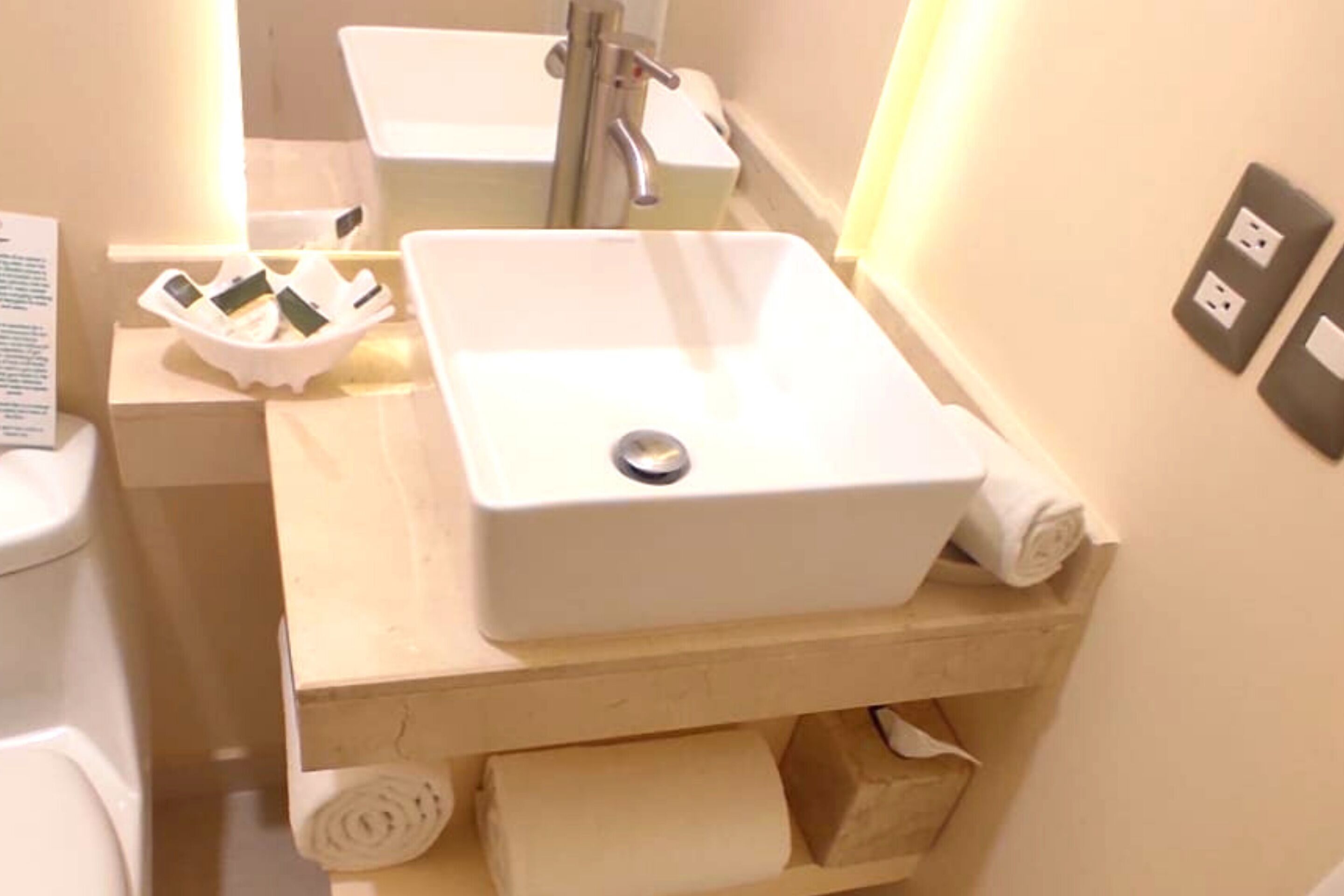 Casa de banho com lavatório branco, espelho, sanita e prateleira com toalhas e artigos de higiene pessoal.
