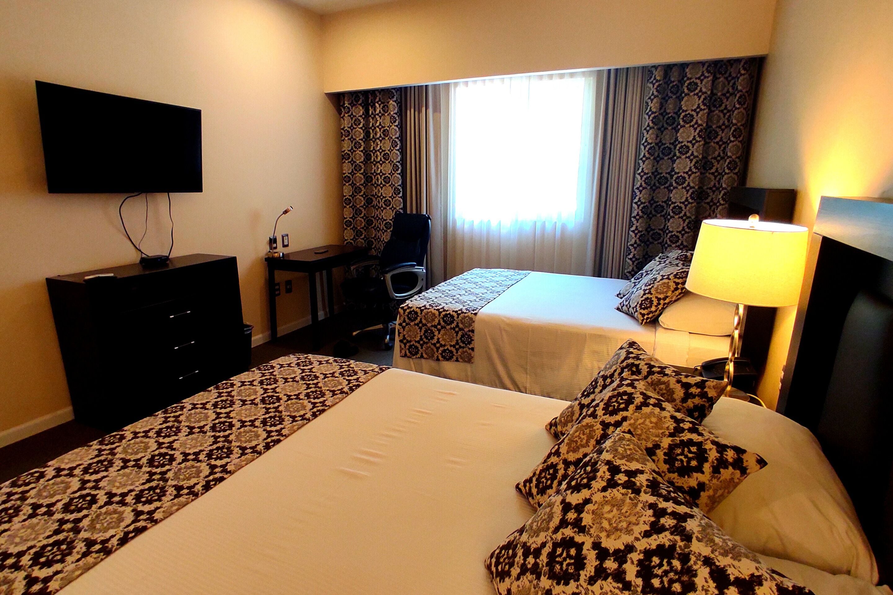 A hotel room with two beds, a desk, a chair, a TV, and a window with curtains.