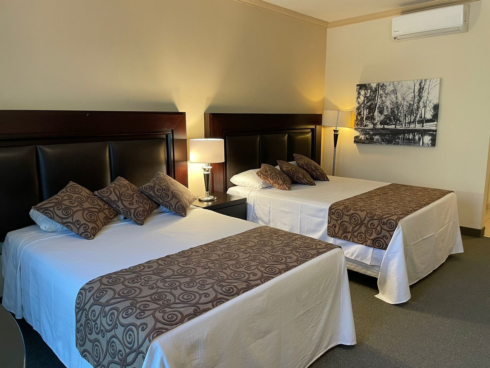 A hotel room with two beds, each with a headboard, a bedside table with a lamp, and a wall-mounted air conditioner.