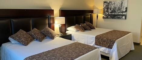 A hotel room with two beds, each with a headboard, a bedside table with a lamp, and a wall-mounted air conditioner.
