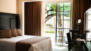 A hotel room with a bed, a desk with a mirror, and a view of a tree through a window. - Hotel Real del Bosque Golf and Spa (Tula de Allende)