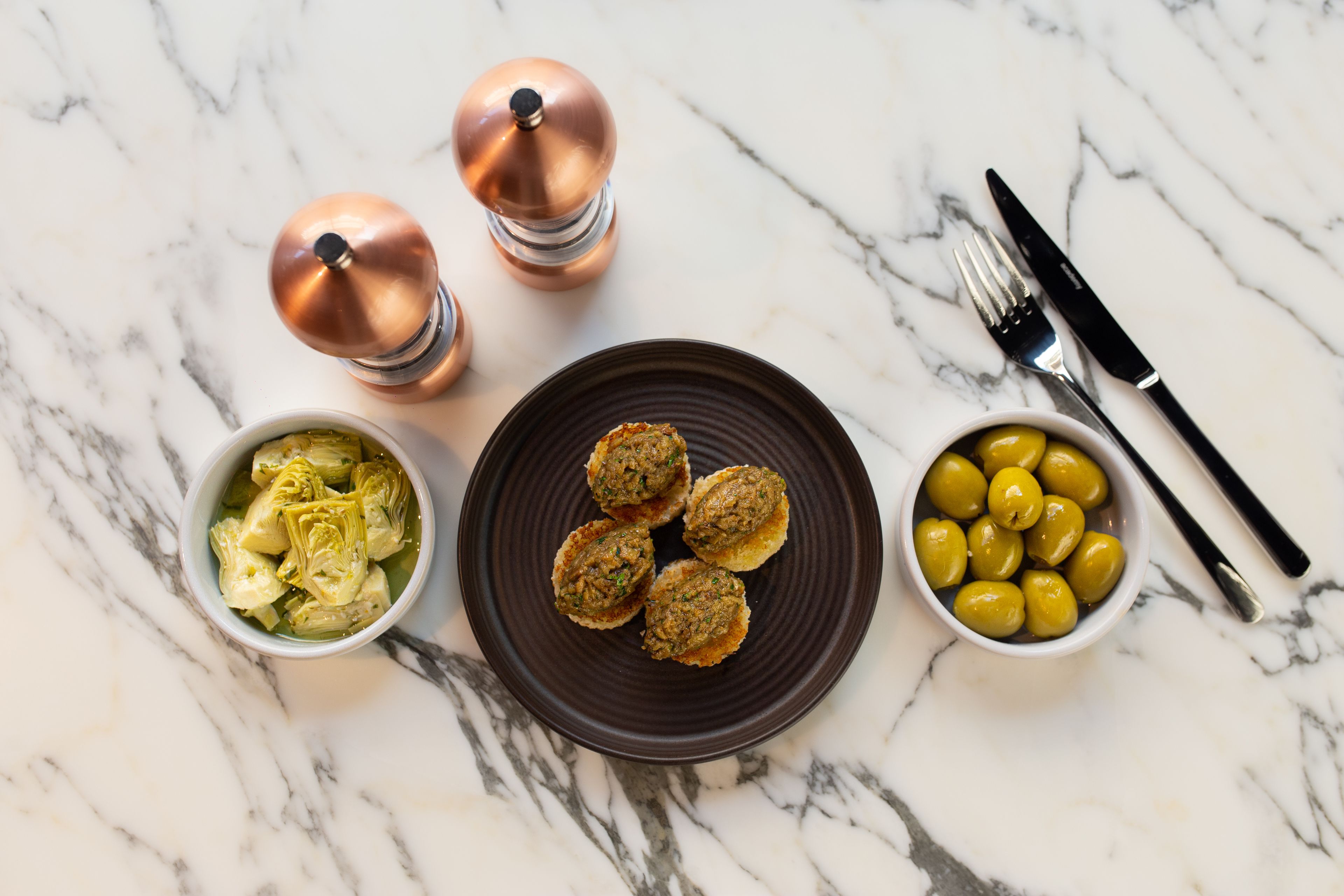 Una mesa de mármol con una porción de falafel, un cuenco de alcachofas y un cuenco de aceitunas.