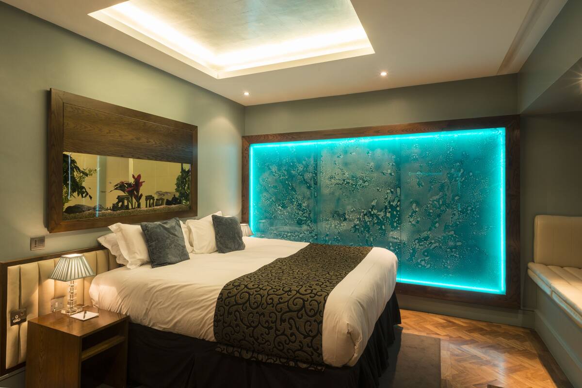 A modern bedroom with a large bed, a nightstand with a lamp, a wall-mounted mirror, and a unique illuminated wall feature.