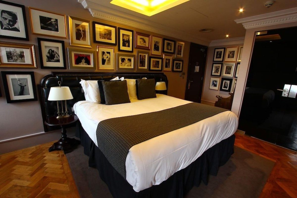 A hotel room with a large bed, a nightstand, a lamp, and a wall adorned with framed pictures.