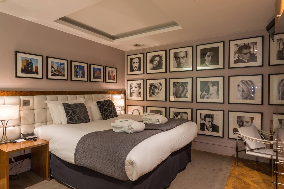 A hotel room with a large bed, a bedside table with a lamp, a chair, and a wall adorned with framed pictures.