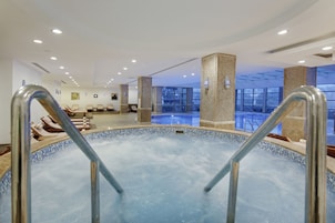 Una piscina interior moderna con agua azul cristalina, rodeada de sillones de descanso y una zona de recepción con un amplio ventanal.